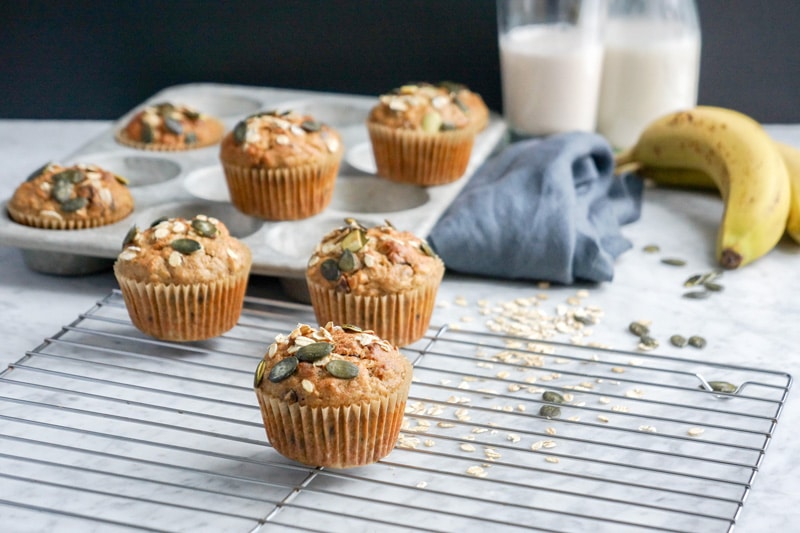 Front view of Multigrain Banana Chocolate Chip Muffins in a muffin pan and on a cooling rack on top of white marble table with fresh bananas, 2 glasses of milk and a grey napkin in the blurry background