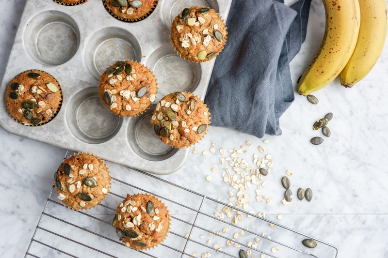 Aerial view of Multigrain Banana Chocolate Chip Muffins in a muffin pan and on a cooling rack on top of white marble table with fresh bananas and a grey napkin on the side