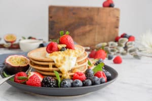 black plate with a stack of small pancakes, fresh berries on the side, and a dollop of yogurt and passion fruit dripping on the side of the pancakes