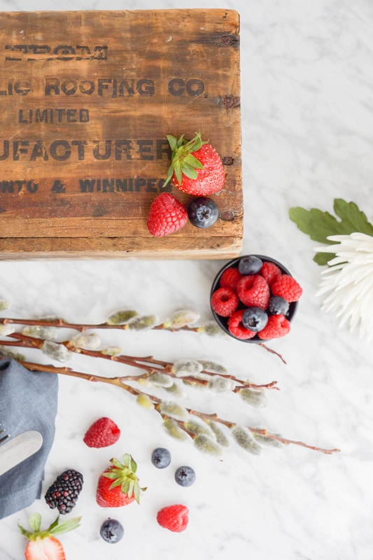 fresh berries in a small black bowl and a wooden box on top of a marble counter with flowers