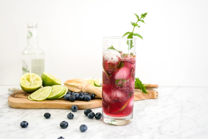 front view of a tall drink with fresh blueberries and limes on a white marble table