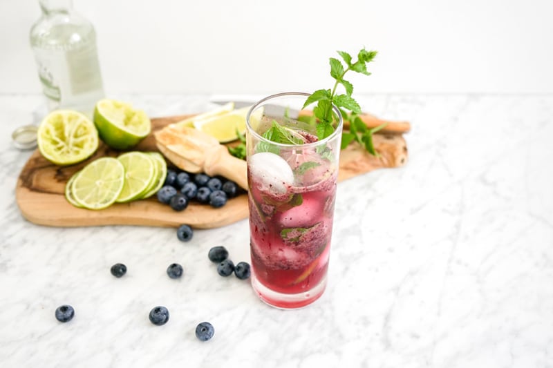 45 degree angle view of a blueberry mojito with blueberries, limes and mint around