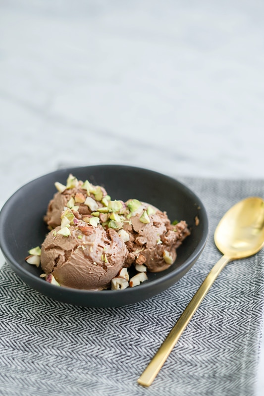 vertical front view of a black bowl with chocolate ice cream in it and sprinkled pistachios and hazelnuts on top