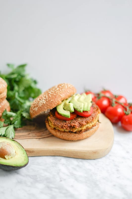 front view of sweet potato chickpea burger on a wood board with tomatoes in the background and an avocado in the foreground