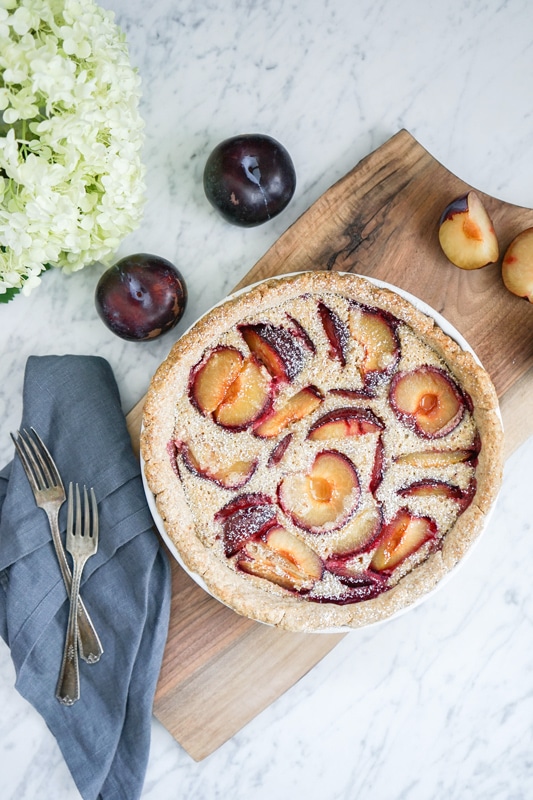 aerial view of a French plum tart with fresh plums on the side and a big white hydrangea bloom 