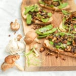 horizontal view of Roasted Mushroom, Onion and Snow Pea Crostini on top of a wood board with fresh mushrooms on the side