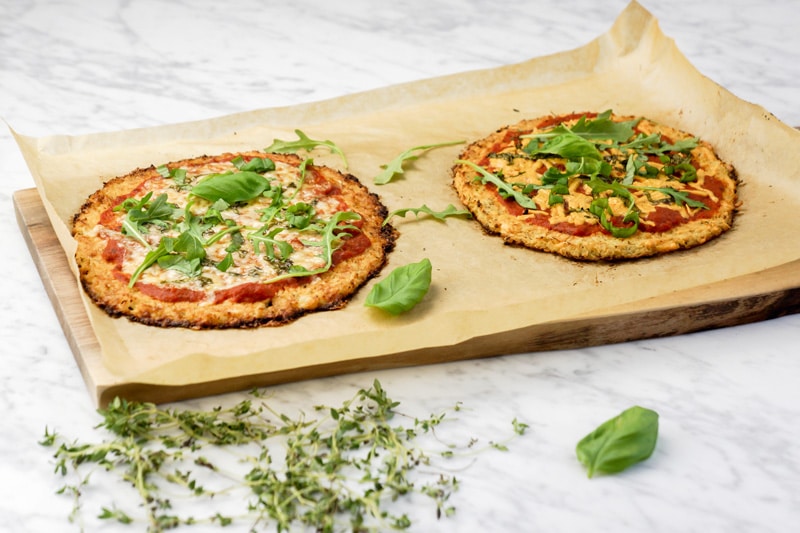 view of 2 Cauliflower Crust Pizzas with fresh basil leaves and baby arugula on top of parchment paper on a cutting board, on a white marble table with fresh thyme and a basil leaf in the forefront