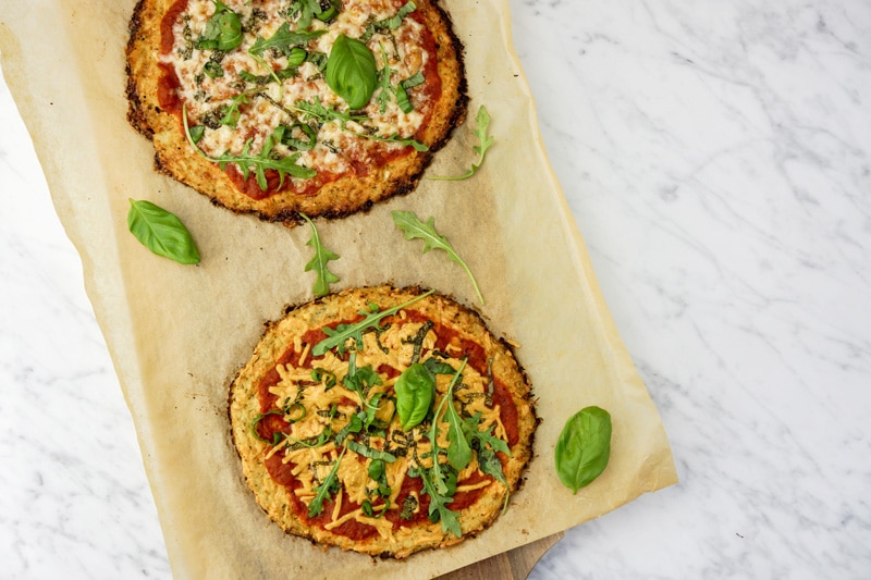 aerial view of 2 Cauliflower Crust Pizzas with fresh basil leaves and baby arugula on top of parchment paper on a cutting board, on a white marble table
