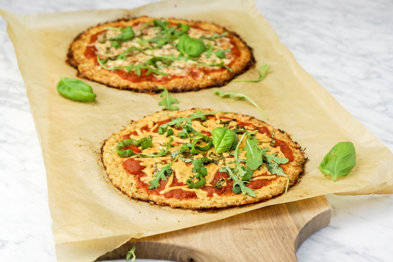 portrait view of 2 Cauliflower Crust Pizzas with fresh basil leaves and baby arugula on top of parchment paper on a cutting board, on a white marble table