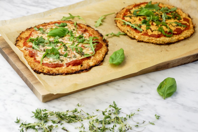 front view of 2 Cauliflower Crust Pizzas with fresh basil leaves and baby arugula on top of parchment paper on a cutting board, on a white marble table with fresh thyme on the forefront