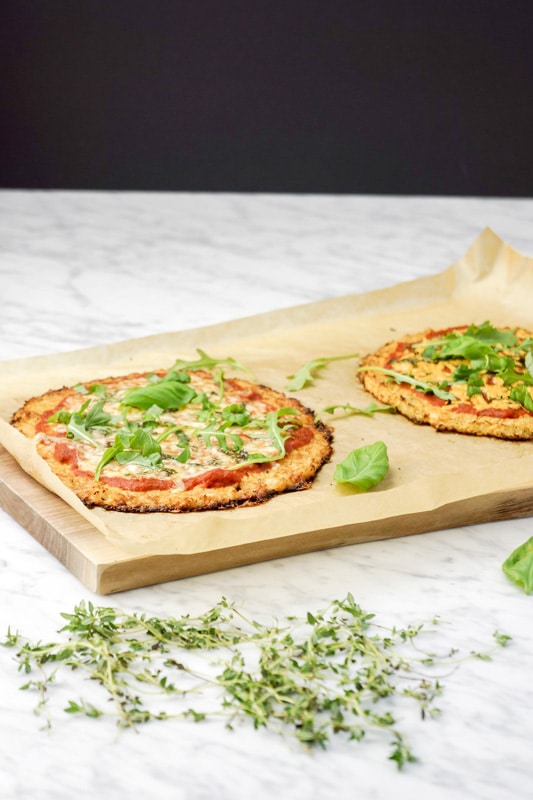 front view of 2 Cauliflower Crust Pizzas with fresh basil leaves and baby arugula on top of parchment paper on a cutting board, on a white marble table with fresh thyme on the forefront and a black background