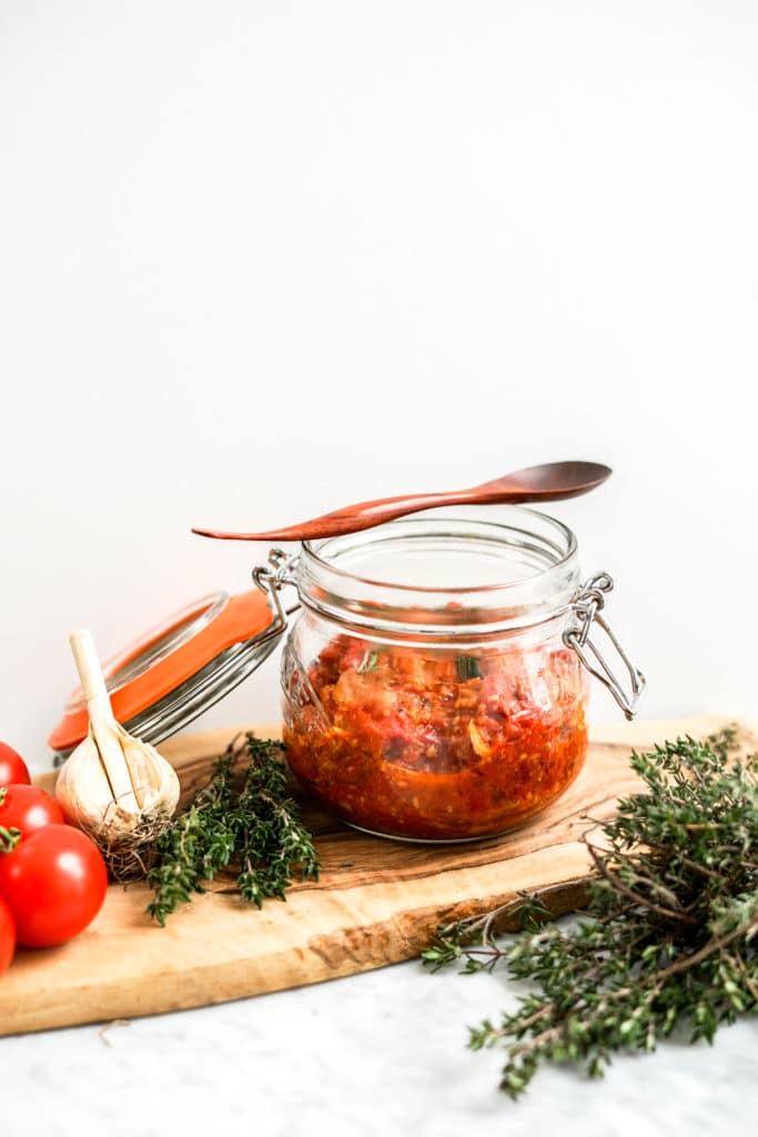 Tomato and garlic confit in a glass canning jar with fresh tomatoes and herbs around it