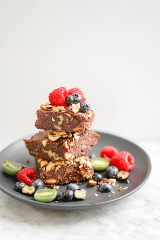 3 black bean brownies piled on top of a black plate with fresh berries around