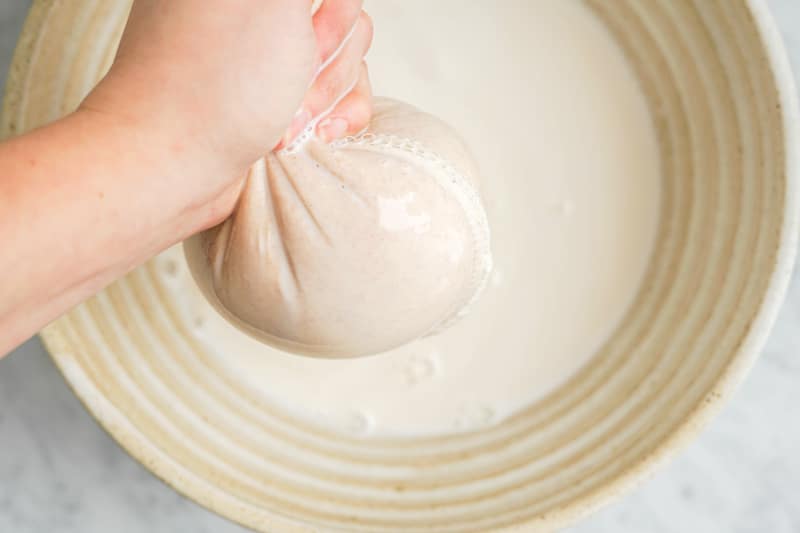 a hand squeezing a nut milk bag over a large ceramic bowl