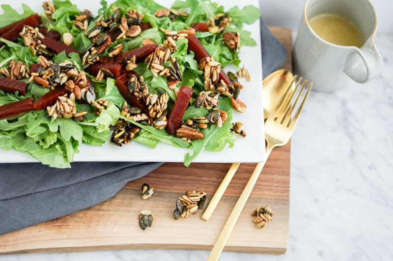 close-up view of a green salad with chopped beets and candied nuts and seeds with golden utensils on the right