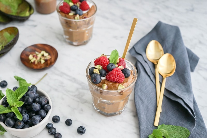 Close-up of a glass filled with chocolate avocado mousse and fresh berries on top, with a bowl of blueberries on the left and 2 golden spoons on a blue napkin on the right
