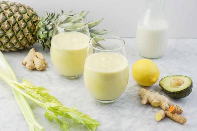 front view of 2 glasses with a light yellow colour smoothie. celery branches on the left side, a pineapple in the background, some fresh ginger and turmeric root, a lemon and avocado on the right, and a glass container of nut milk in the back