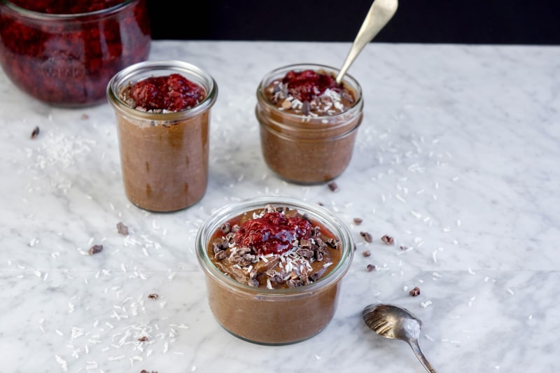 front view of 3 small jars of chocolate pudding and strawberry compote on top, with shredded coconut pieces and cacao nibs scattered around, on a white marble table