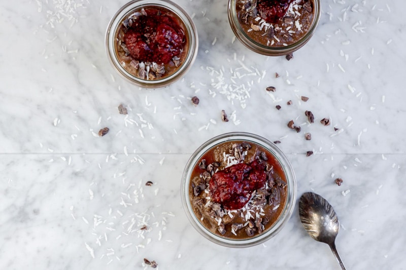 aerial view of 3 small jars of chocolate pudding and strawberry compote on top, on a white marble table, with shredded coconut pieces and cacao nibs scattered around