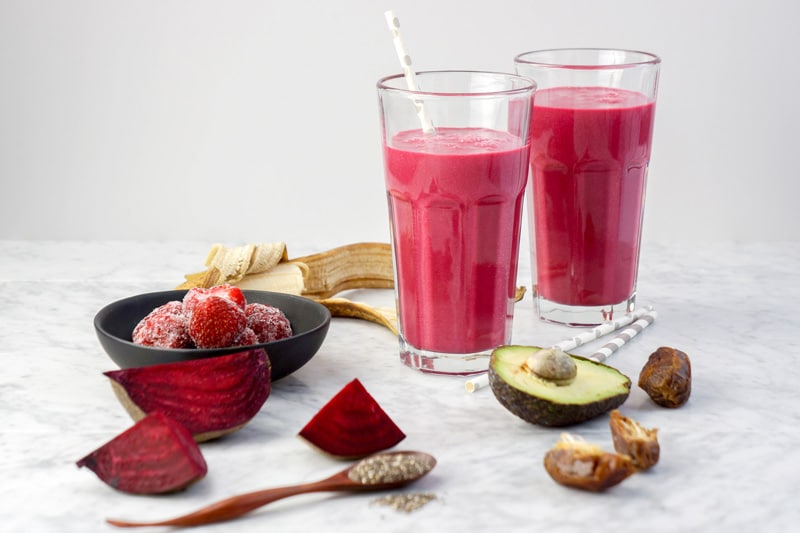 2 glasses of deep pink smoothie, with beet roots, avocado, dates, strawberries, and a banana around, on a white background and white marble tabletop