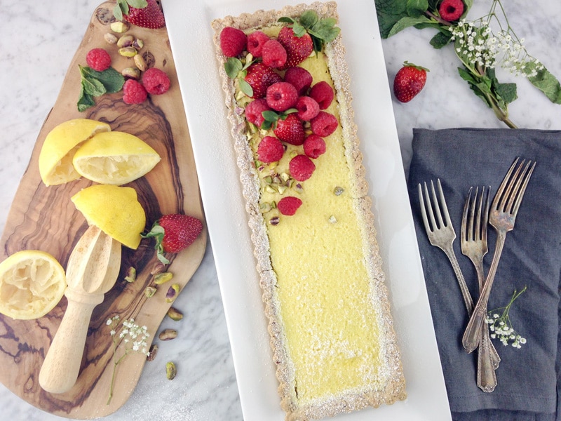 horizontal aerial view of a rectangular lemon tart on a white plate, with raspberries and strawberries on top, and some squeezed lemon halves and fresh berries on a wood board on the left side and forks on a grey napkin on the right side