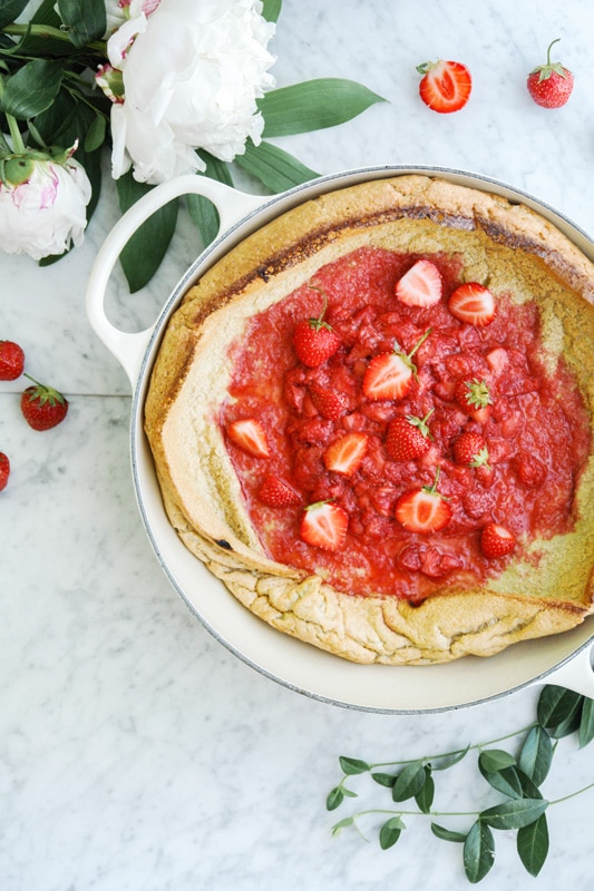 horizontal aerial view of a Baby Dutch Pancake with a strawberry compote in the middle, in a beige enamelled cast iron pan surrounded by fresh peonies and strawberries