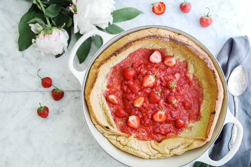 landscape aerial view of a Baby Dutch Pancake with a strawberry compote in the middle, in a beige enamelled cast iron pan surrounded by fresh peonies, strawberries, a grey napkin and an antique spoon