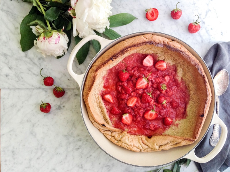 landscape aerial view of a Baby Dutch Pancake with a strawberry compote in the middle, in a beige enamelled cast iron pan surrounded by fresh peonies and strawberries