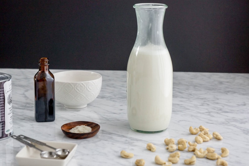 front horizontal view of a bottle of milk with cashews on one side, a small dish of salt, a can of maple syrup, a bottle of vanilla and a white bowl in the background, on a white marble table and a black background