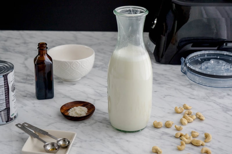 front view of a bottle of milk with cashews on one side, a small dish of salt, a bottle of vanilla and a can of maple syrup on the side and a small white bowl in the background, on a white marble table, with a partial view of a blender in the right corner