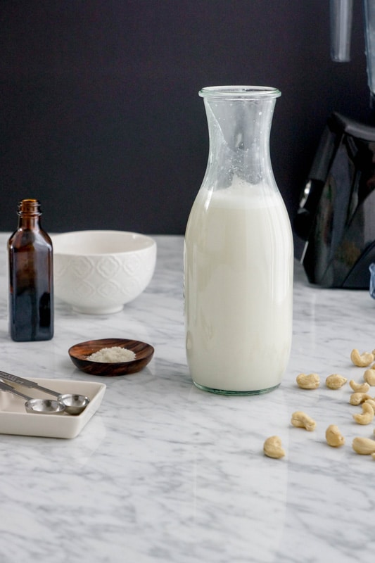 front horizontal view of a bottle of milk with cashews on one side, a small dish of salt, a bottle of vanilla and a white bowl in the background, on a white marble table and a black background